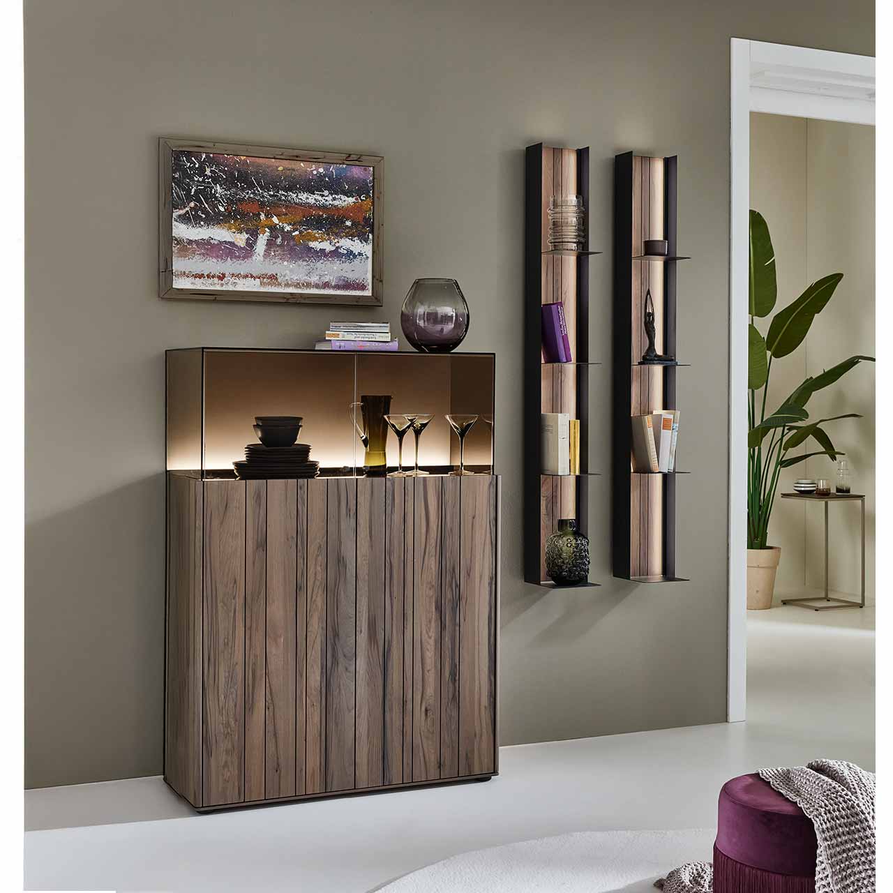 Modernes Highboard aus dunkelbraunem Holz mit sichtbarer Maserung, oben mit aufgesetztem Glaskasten und beleuchteter Rückwand in sandfarbenem Lack. Daneben zwei vertikale Wandregale aus Metall mit je vier Ablageböden – vor dunkelbeiger Wand
