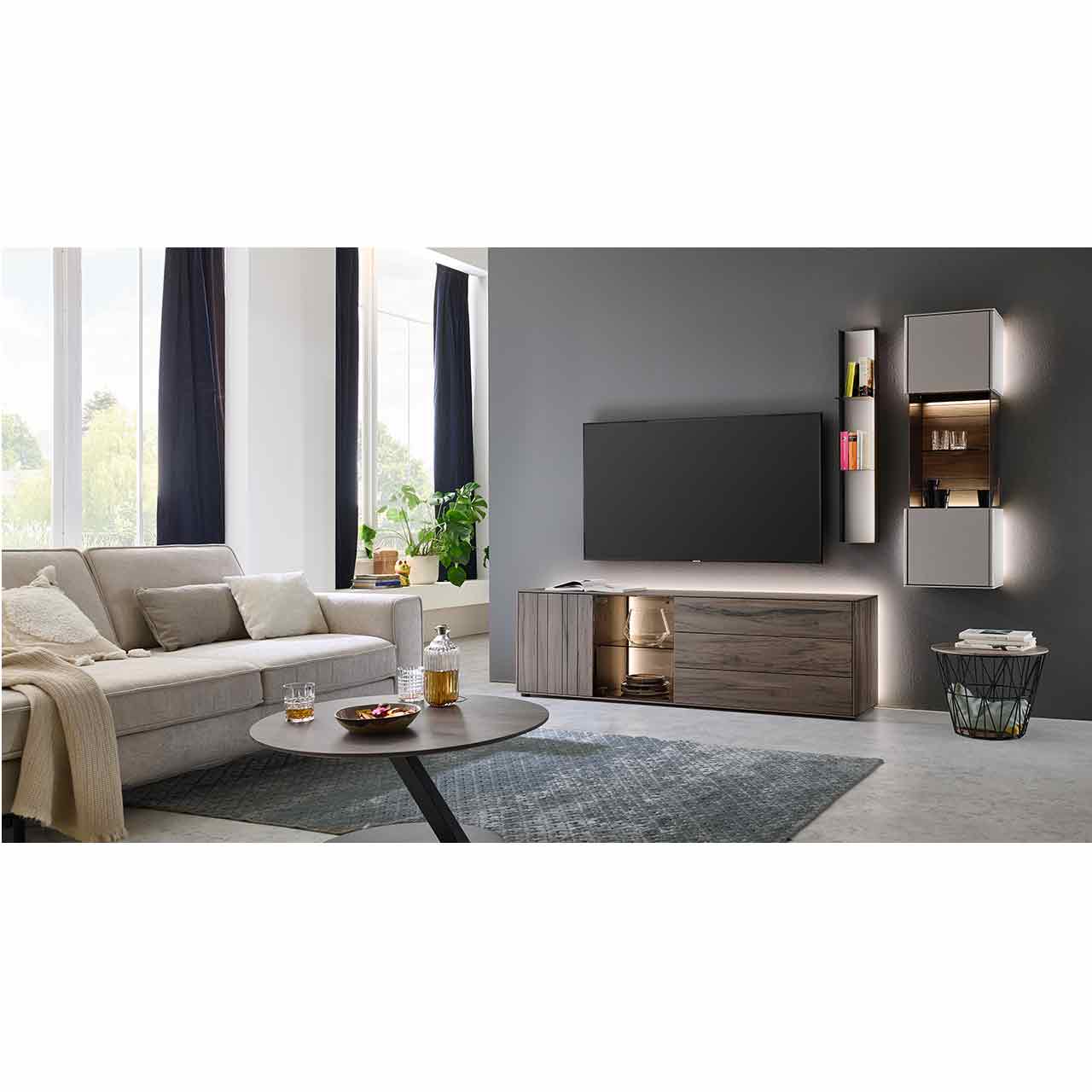 Modernes Wohnzimmer mit dreiteiliger Wohnwand bestehend aus dunkelbraunem Holzlowboard mit Glasausschnitt, Metallregal mit beleuchteter Lackfront und Metallböden und einer Hängevitrine in Lack sand vor rundem Couchtisch und heller Couch.