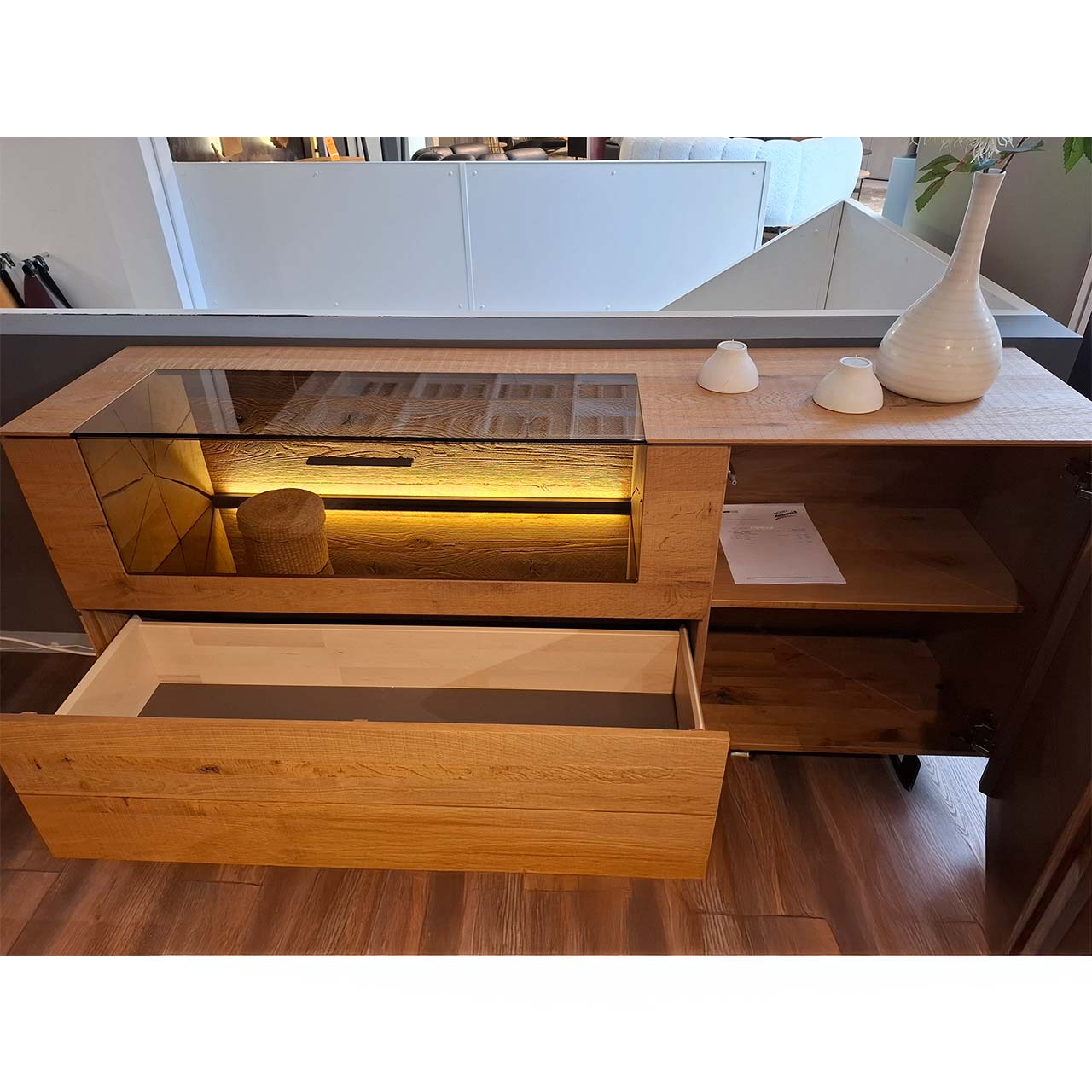 Modernes Sideboard aus massiver Kerneiche Umato auf zwei Metallkufen mit beleuchtetem Glasausschnitt, offenem Schubkasten und geöffneter Tür.
