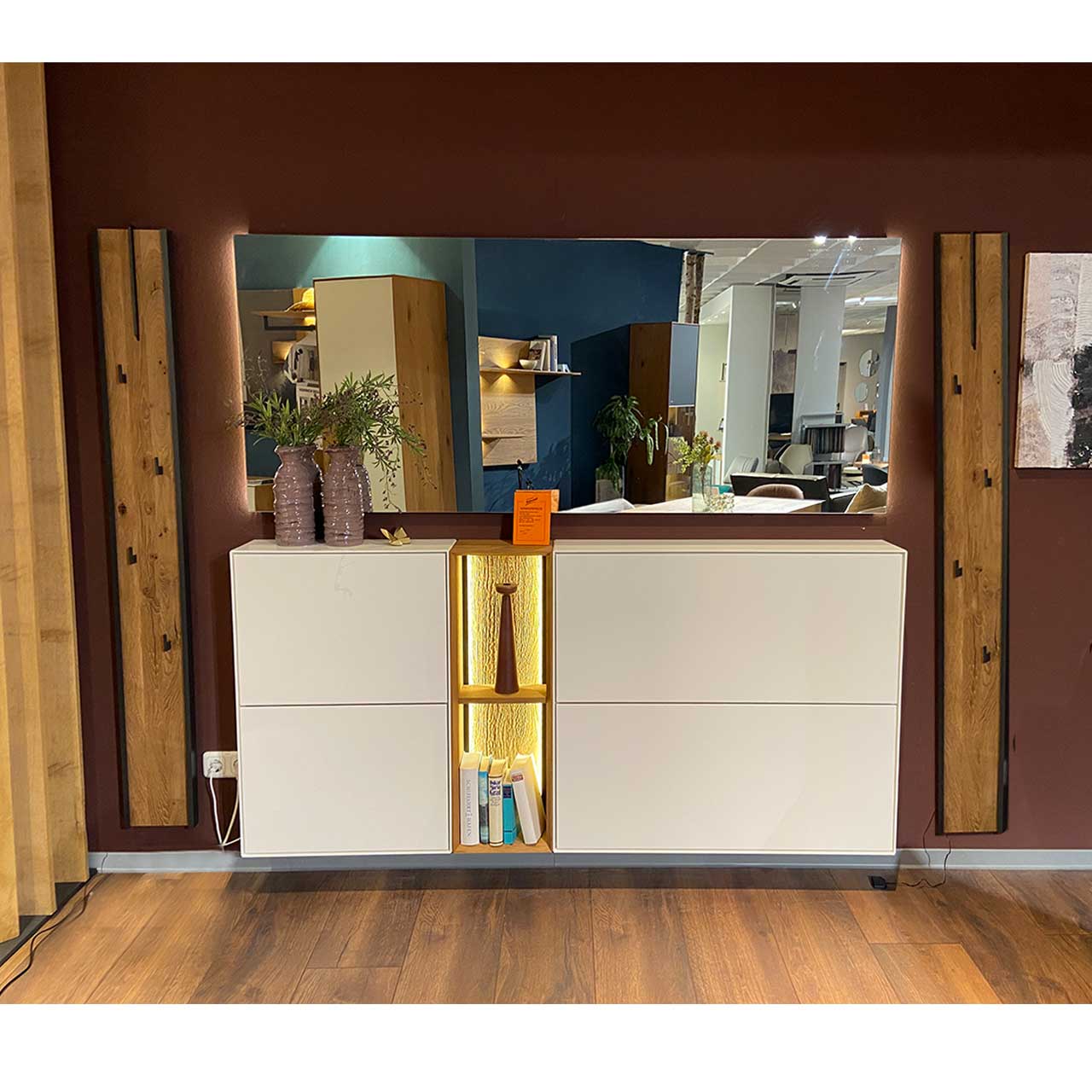 Hartmann SALE Möbel - VELKO Garderobenkombination 7240G Nr. V100 inkl. Beleuchtung / Bild bearbeit aus Ausstellung Moderne Garderobenkombination mit zwei weißen, hängenden Schuhkippern, mittigem offenem Regalelement mit Beleuchtung, einem breiten Wandspiegel und zwei vertikalen Garderobenpaneelen aus massiver Kerneiche mit schwarzen Metallhaken.