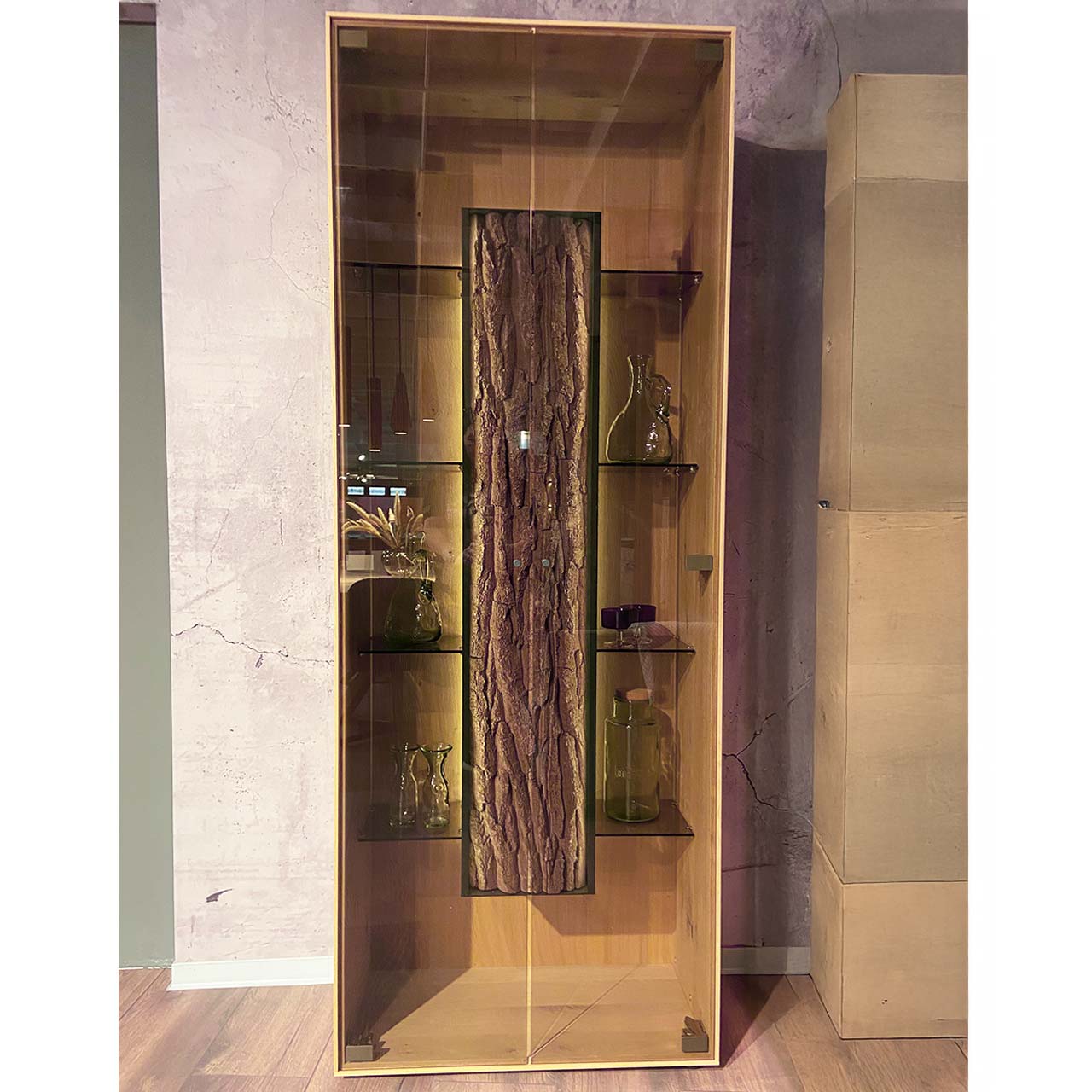 Hartmann SALE Möbel - RUNA Standvitrine 8410-0081 - inkl. Beleuchtung / Originalbild Ausstellung Moderne Standvitrine aus massiver Kerneiche Natur mit zwei Glastüren, innenliegenden Glasböden und mittigem Korpus mit beleuchteter Baumrinden-Applikation.
