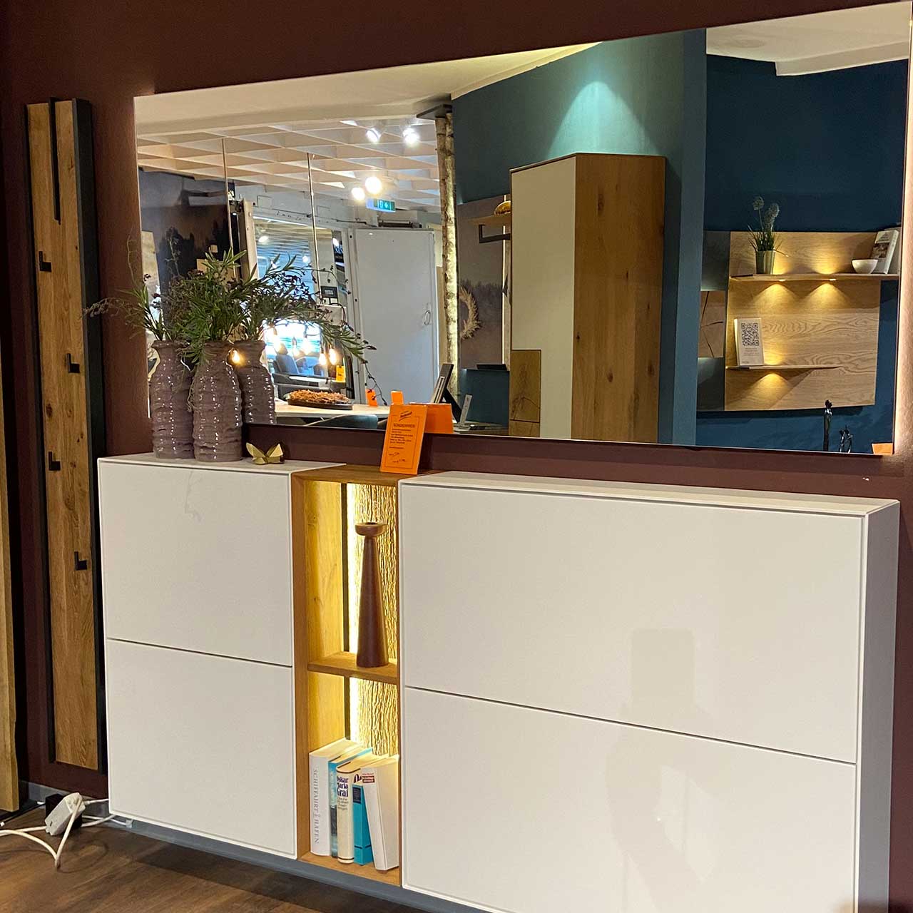 Moderne Garderobenkombination mit zwei weißen, hängenden Schuhkippern, mittigem offenem Regalelement mit Beleuchtung, einem breiten Wandspiegel und links vertikales Garderobenpaneelen aus massiver Kerneiche mit schwarzen Metallhaken.