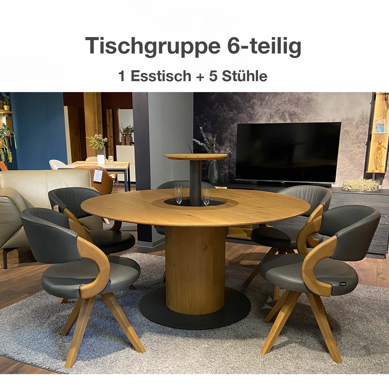 Hartmann SALE Möbel - NATURZEIT Esstischgruppe 6-teilig 7100E-1557 und 1637 / Originalbild aus Ausstellung Moderne Esstischgruppe aus massiver Kerneiche Umato, bestehend aus einem runden Esstisch mit mittigem, offenem Flaschenfach in der Tischplatte. Um den Tisch stehen fünf drehbare Stühle mit Holzlehne und Holzgestell, bezogen mit grauem Leder.
