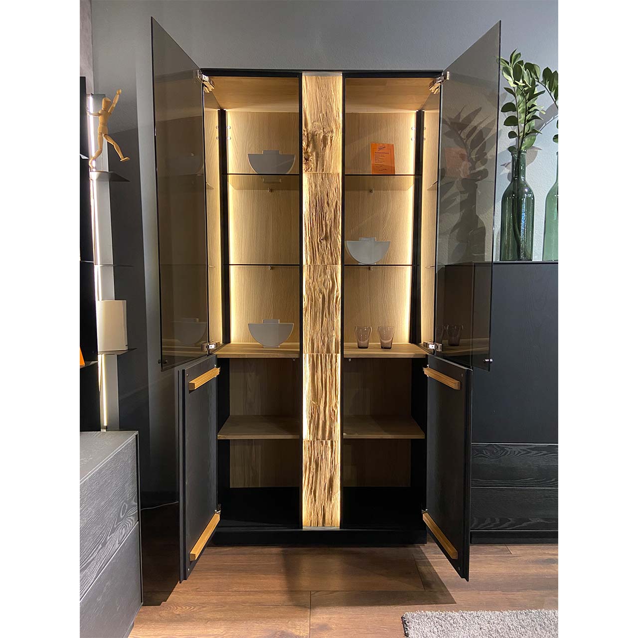 Moderne Standvitrine aus anthrazitfarbener Kernesche mit vier geöffneten Holz- und Glastüren, beleuchteter Rückwand, Glasböden und mittigem Spaltholz-Akzent.