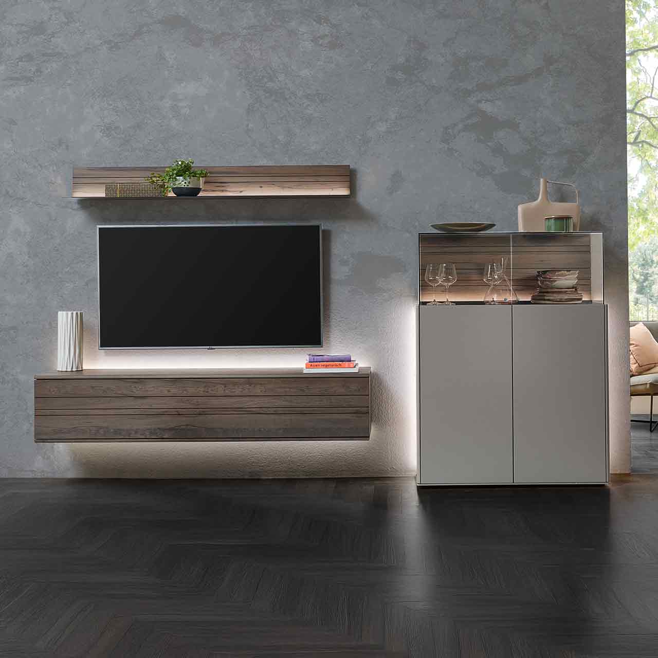 Hartmann AUDRA 5610 - Wohnwand Nr. V34 - dreiteilig aus Altholzbuche Uyaa und Lack Sand Modernes Wohnzimmer mit dreiteiliger Wohnwand bestehend aus Hängelowboard und Wandregal in dunkelbraunem Holz mit Maserung, daneben Highboard in Lack Sand mit aufgesetztem Glaskasten.