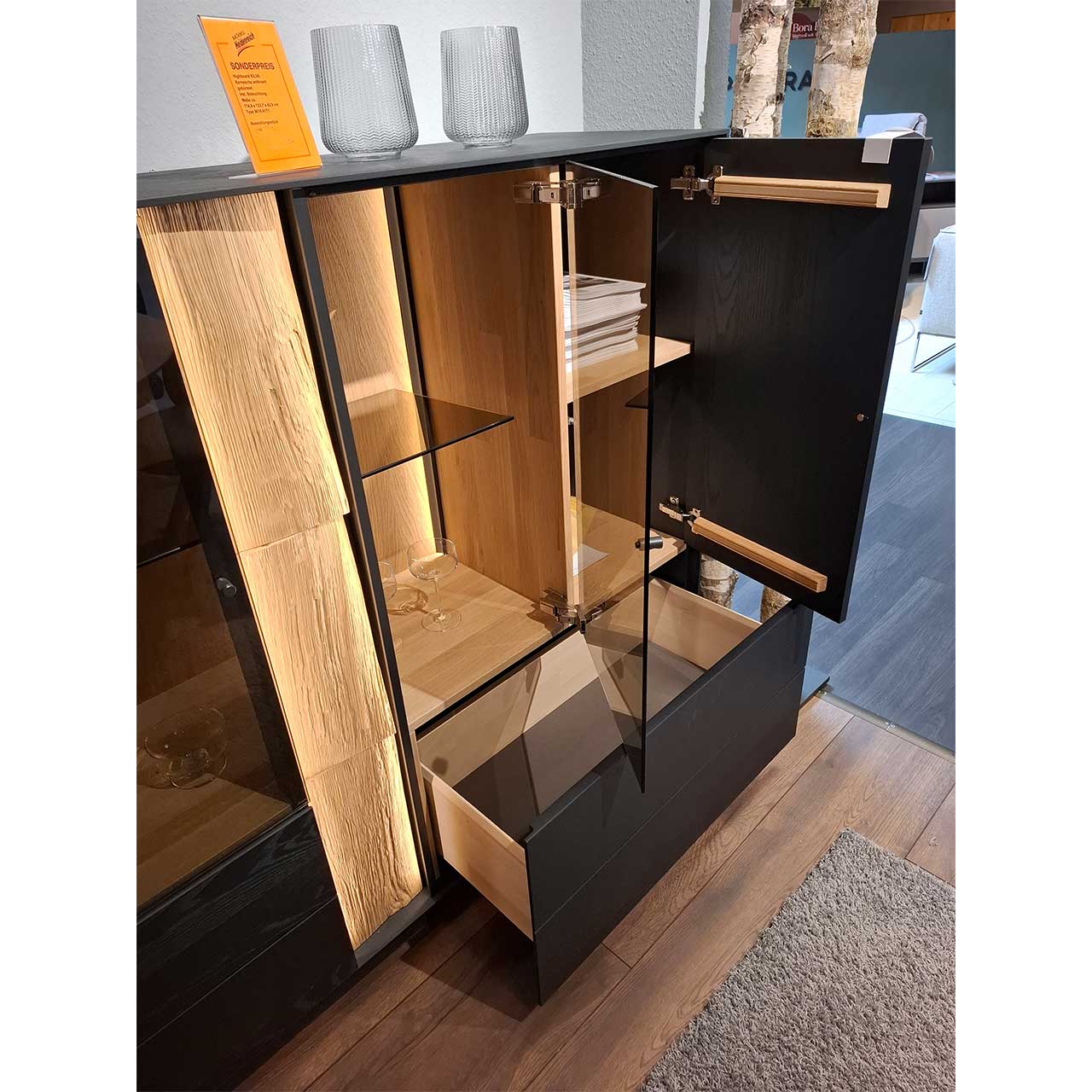 Bildausschnitt eines Highboards aus anthrazitfarbener Kernesche mit geöffneter Glas- und Holztür, offenem Schubkasten, beleuchteter Rückwand, Glasböden und vertikalem Spaltholz-Akzent