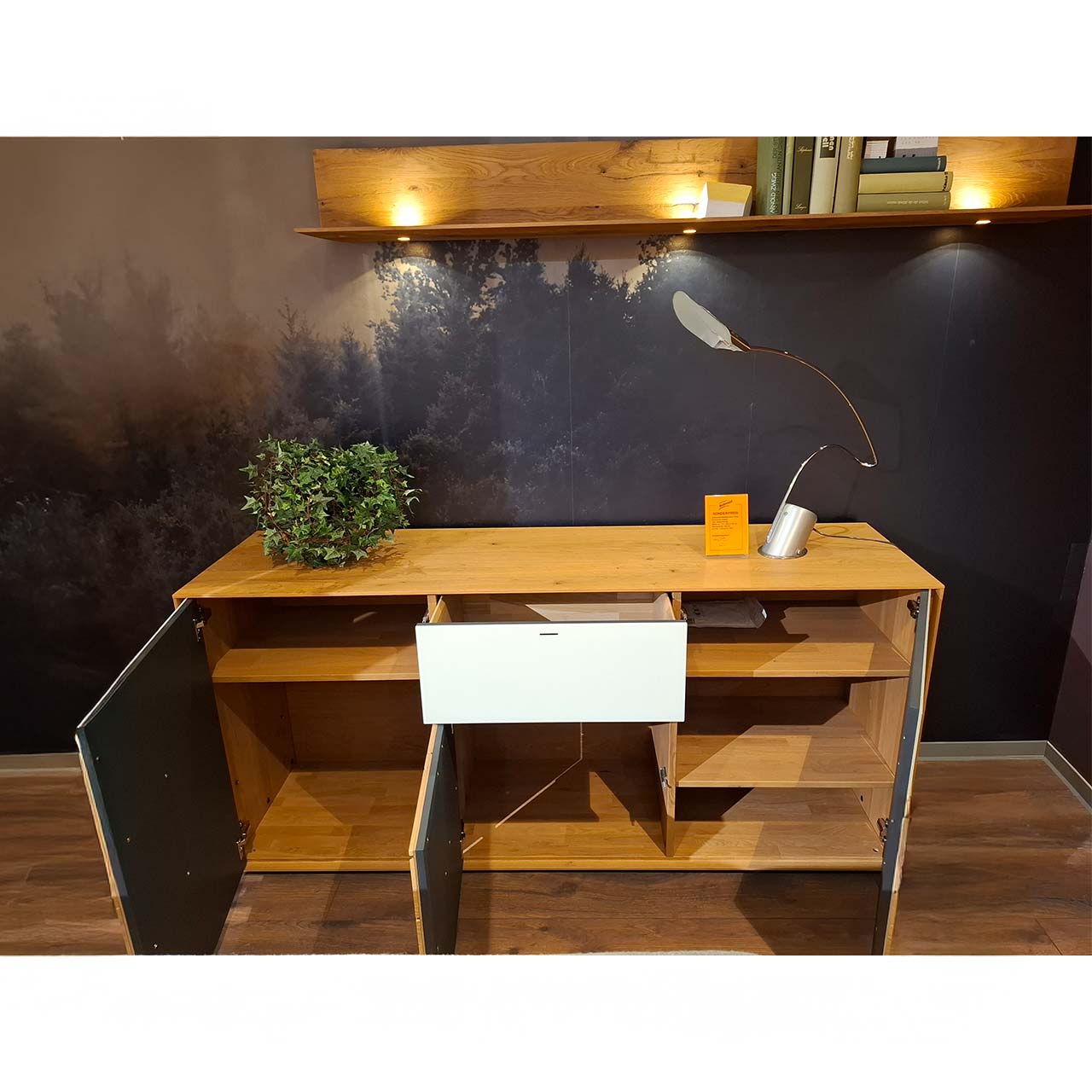 Möbel ZweierSet: Modernes Sideboard aus massiver Kerneiche, rechts und links mit geöffneten Türen und sichtbaren Holzböden, mittig mit offenem Schubkasten und offener Tür. Darüber hängend ein langes Wandpaneel mit Spotbeleuchtung.