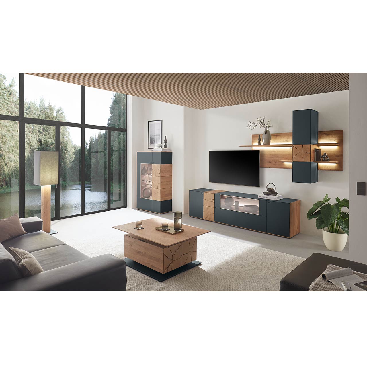 Hartmann CAYA - Highboard 7170-7081A Moderne Wohnwand aus massiver Kerneiche mit anthrazitfarbenen Fronten, mit Lowboard mit Glasausschnitt sowie Wandpaneel mit Regalböden und Hängeschrank. Links ein Highboard mit Glasfront und Hirnholzscheiben. Im Vordergrund der passende Couchtisch.