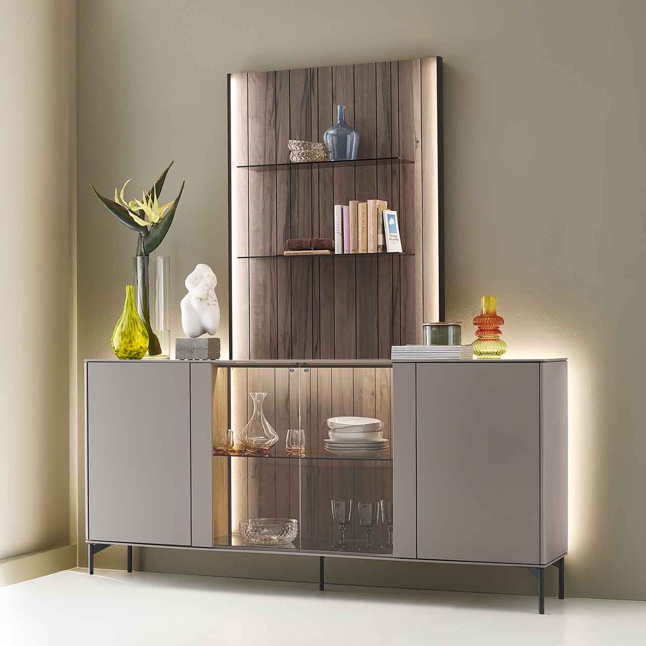 Modernes Massivholz-Sideboard auf Metallfüßen mit zwei Türen rechts und links lackiert in Sandfarben, mittig mit Glasausschnitt und Glasboden vor beleuchteter Holzrückwand in edler, dunkelbrauner Altholzbuche Uyaa, darüber Holzpaneel mit Glasböden.