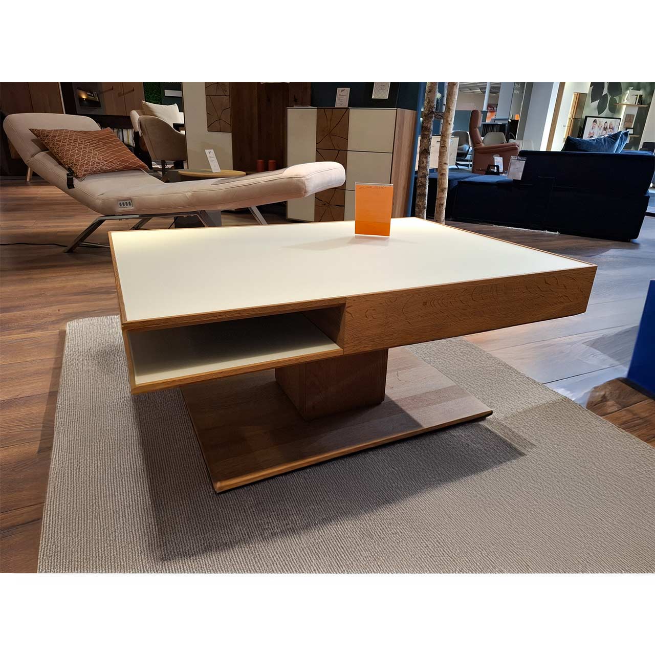 Moderner, rechteckiger Couchtisch aus massiver Kerneiche Umato. Dicke Tischplatte in Mattglas weiß mit integriertem Schubkasten und seitlichem Ablagefach, auf massiver, rechteckiger Holzsäule und Bodenplatte stehend.
