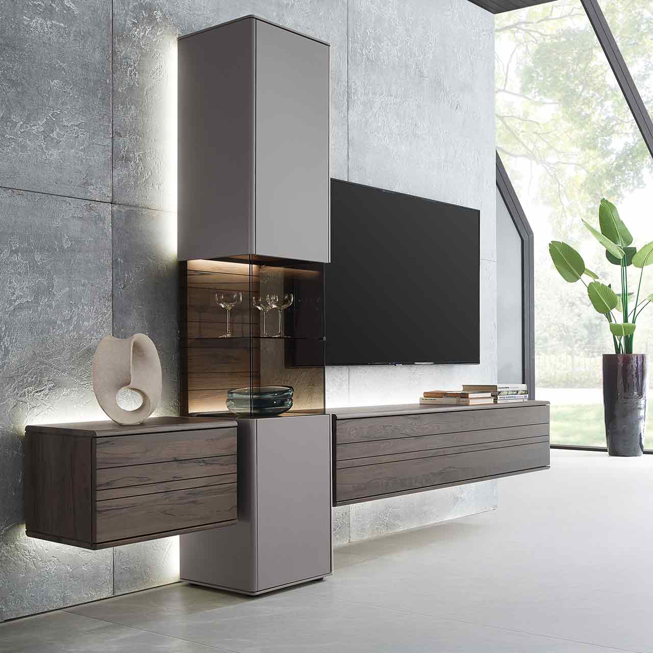 Hartmann AUDRA 5610 - Wohnwand Nr. V20 - dreiteilig aus Altholzbuche Uyaa und Lack Sand Moderne, dreiteilige Massivholz-Wohnwand bestehend aus einer Standvitrine mit Fronten in Lack Sand und beleuchtetem Glasausschnitt, rechts und links davon zwei Hängelowboards in dunkelbraunem Holz mit sichtbarer Maserung.