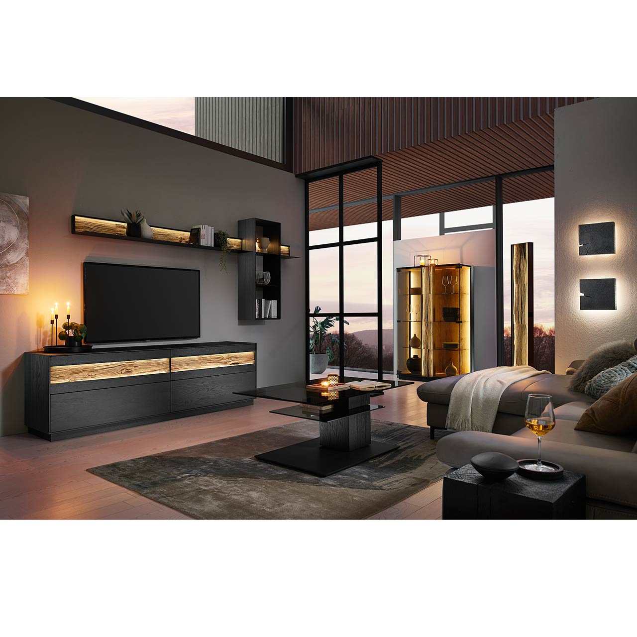 Milieubild Wohnzimmer - Hartmann NATURWERKE - Couchtisch 9600C-1405 Wohnzimmer mit moderner Wohnwand in Kernesche anthrazit: Lowboard, Wandpaneel und Wandregal. Rechts das passende Highboard mit Glasfront. Im Vordergrund ein Couchtisch mit rechteckiger Glasplatte auf massiver, dunkler Holzsäule.