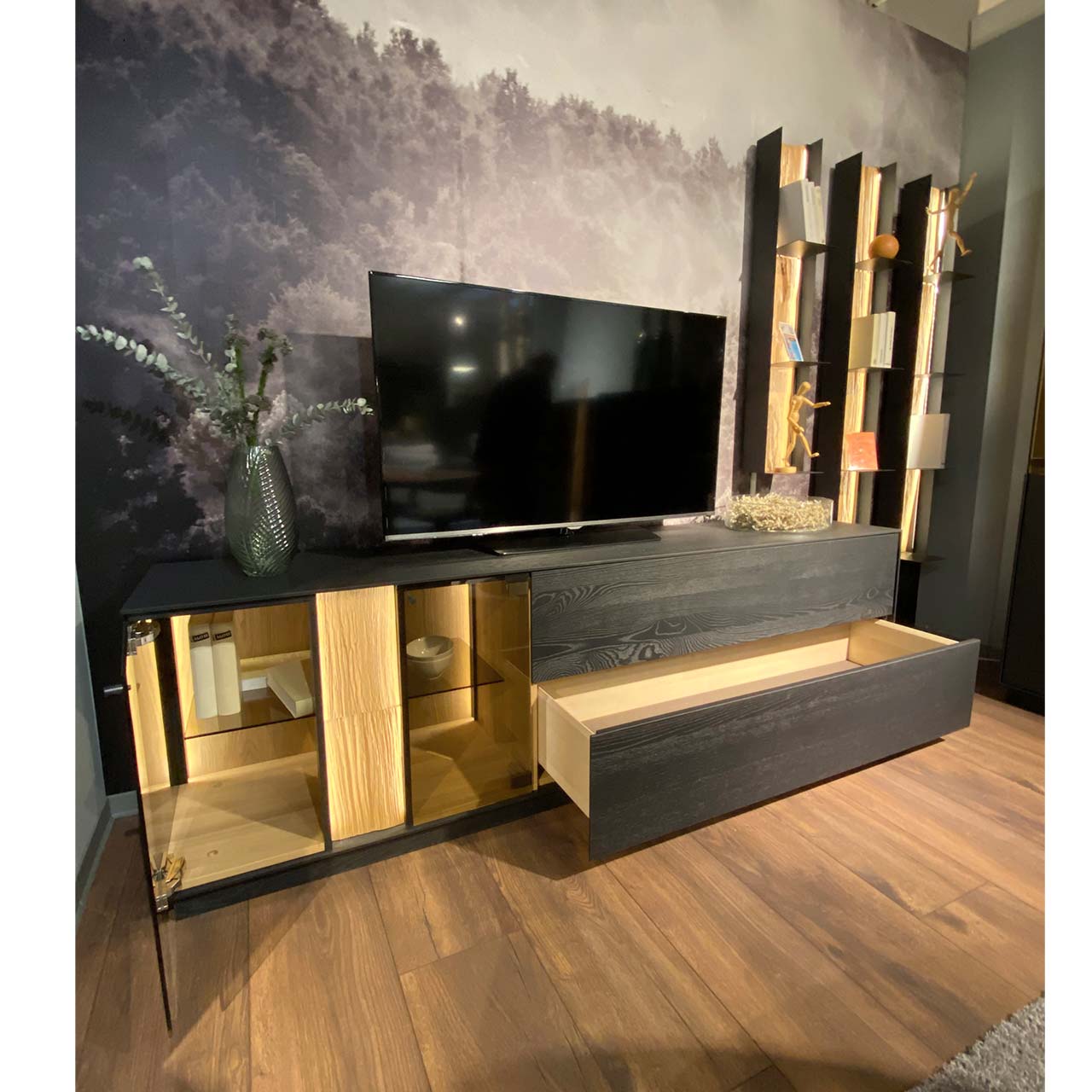 Moderne vierteilige Massivholz Wohnwand aus anthrazitfarbener Kernesche und hellem Spaltholz: Lowboard mit Glasausschnitt, Klappe und geöffnetem Schubkasten, drei Metallregale mit Ablageböden und beleuchteter Spaltholzrückwand.