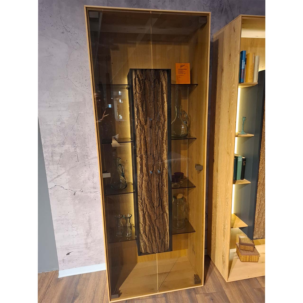 Moderne Standvitrine aus massiver Kerneiche Natur mit zwei Glastüren, innenliegenden Glasböden und mittigem Korpus mit beleuchteter Baumrinden-Applikation.