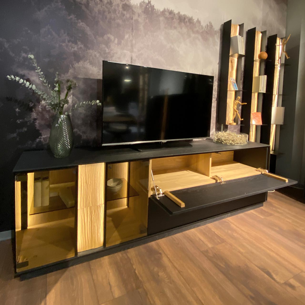 Moderne vierteilige Massivholz Wohnwand aus anthrazitfarbener Kernesche und hellem Spaltholz: Lowboard mit Glasausschnitt, Schubkasten und geöffneter Klappe, drei Metallregale mit Ablageböden und beleuchteter Spaltholzrückwand.