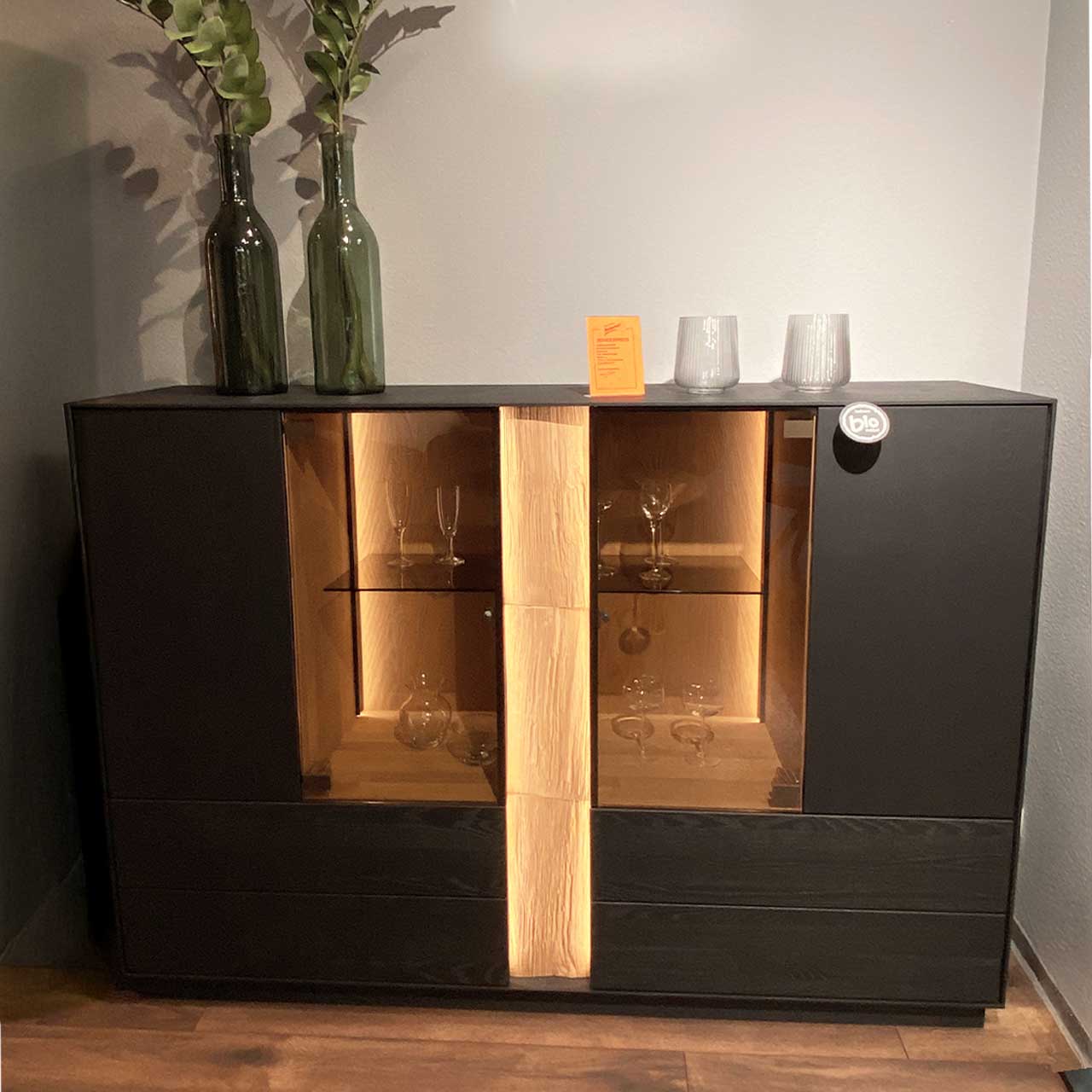 Hartmann SALE Möbel - KILVA Highboard 9610-6171 - inkl. Beleuchtung / Originalbild Ausstellung Modernes Highboard aus anthrazitfarbener Kernesche mit mittigem beleuchtetem Glasausschnitt und vertikalem Spaltholz-Akzent, innen mit Glaseinlegeböden. Links und rechts geschlossene Fronten.
