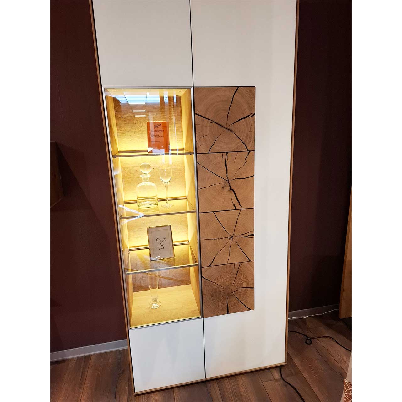 Hartmann SALE Möbel - CAYA Standvitrine 7170-0097W re. - inkl. Beleuchtung / Originalbild Ausstellung Moderne Standvitrine aus Massivholz mit weißen Mattglasfronten, rechts mit Tür und drei Hirnholzscheiben in heller Kerneiche, links mit Tür und beleuchtetem Glasausschnitt.
